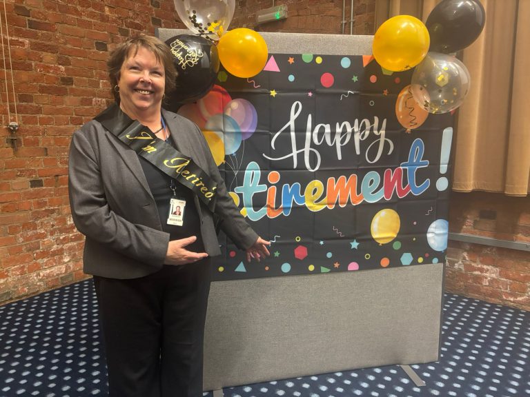 A person wearing a dark suit and a sash that reads “I’m Retired” stands in front of a colorful banner that says “Happy Retirement!” The banner is decorated with multicolored letters, balloons, and confetti designs. There are black, gold, and clear balloons attached to the top corners of the banner. The setting has exposed brick walls and a patterned carpet.