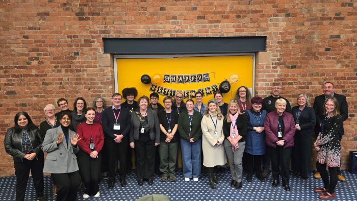 A large group of people standing together in front of a bright yellow wall with a “Happy Retirement” banner and black and gold balloons. The group is dressed in business or smart casual attire, and the background features exposed brick walls and a patterned carpet.