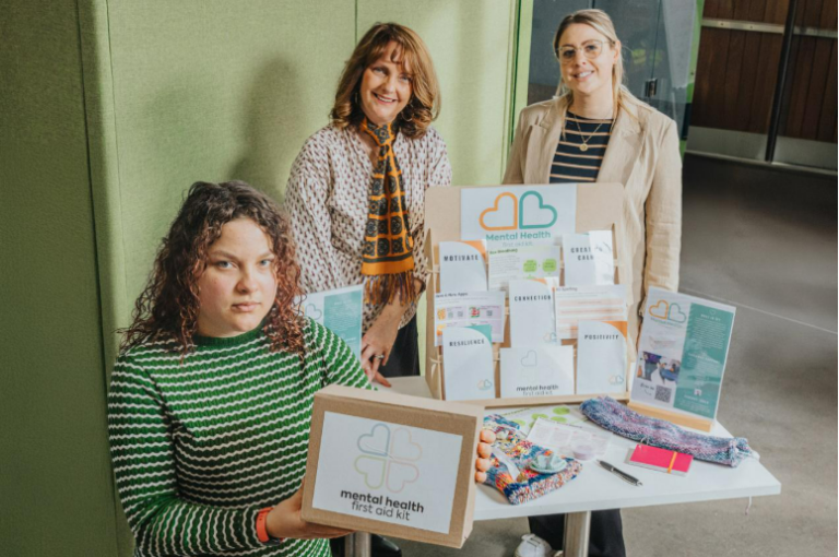A table showcasing multiple mental health first aid kits and related materials. The display includes several labeled envelopes with words like “Motivate,” “Relax & Feel,” “Connection,” “Positivity,” and “Resilience.” There is also a cardboard box with the kit’s logo, an informational stand, a colorful knitted fabric, and a pink notebook. Three individuals are standing behind the table, holding and presenting the kits.