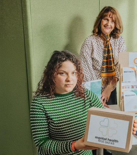 A table showcasing multiple mental health first aid kits and related materials. The display includes several labeled envelopes with words like “Motivate,” “Relax & Feel,” “Connection,” “Positivity,” and “Resilience.” There is also a cardboard box with the kit’s logo, an informational stand, a colorful knitted fabric, and a pink notebook. Three individuals are standing behind the table, holding and presenting the kits.