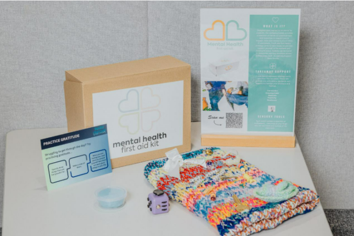 A mental health first aid kit displayed on a white table. The kit includes a cardboard box with a logo featuring a heart-shaped design and the words “mental health first aid kit.” Next to it is an informational stand with details about the kit, a QR code, and a teal section explaining its purpose. Other items on the table include a colorful knitted fabric, a small purple fidget cube, a clear container, and a card titled “Practice Gratitude” with tips for positive thinking.