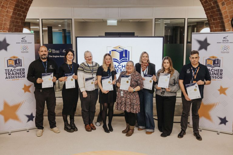 A group of seven people standing in a row holding certificates in front of a large display screen with the logo “Teacher Trainer Assessor” and two banners on either side featuring the same logo and star graphics. The setting appears to be an indoor event space with brick arches and bright lighting.