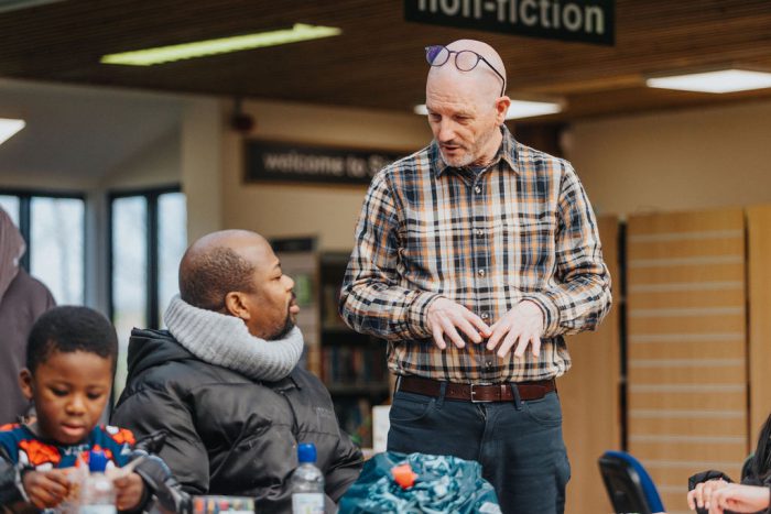 A tutor stands speaking to a small group seated at a table in a library, with bookshelves and signage visible in the background.