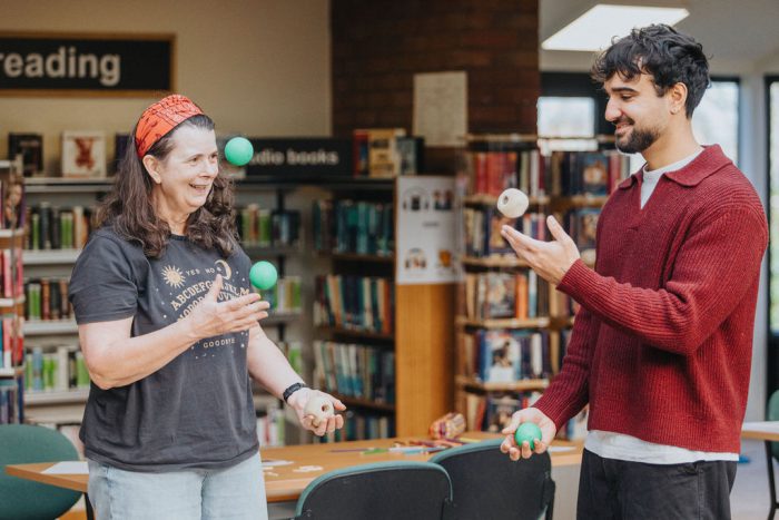 Two people practise juggling with colourful balls inside a library, standing near tables and bookshelves during an activity session at the Adult Learning Open Day at Sinfin Library