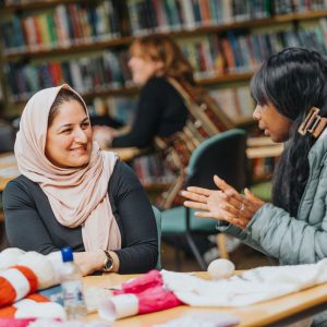 Two adults sit at a library table covered with craft materials, talking and engaging in a creative activity with bookshelves behind them.