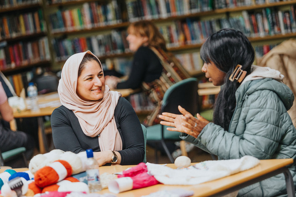 Two adults sit at a library table covered with craft materials, talking and engaging in a creative activity with bookshelves behind them.