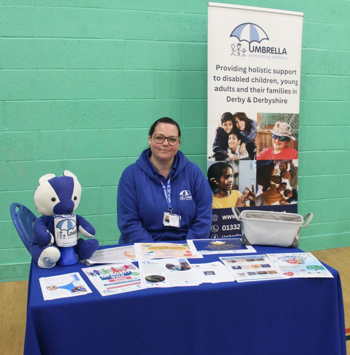 Information stall for Umbrella, a charity supporting disabled children, young adults, and families in Derby and Derbyshire. The table is covered with a blue cloth displaying brochures and leaflets. A large banner behind the table shows the Umbrella logo and tagline. A blue and white teddy bear sits on the table alongside a grey bag.