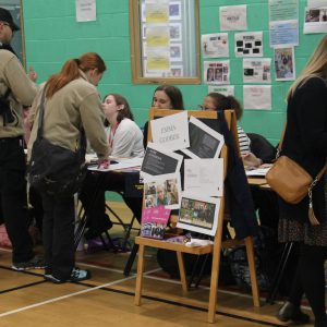 Indoor careers event with tables displaying brochures and information. A chair in the foreground holds a display board titled 'Emma Godber' with details about hobbies and experience. People are standing and interacting with stallholders. The background features green walls and noticeboards with posters and printed materials.