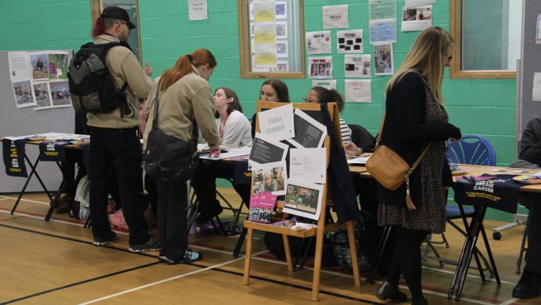 Indoor careers event with tables displaying brochures and information. A chair in the foreground holds a display board titled 'Emma Godber' with details about hobbies and experience. People are standing and interacting with stallholders. The background features green walls and noticeboards with posters and printed materials.