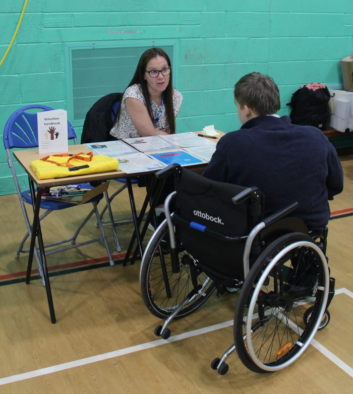 Two individuals seated at a table during an event. One person is in a wheelchair branded 'Ottobock', facing a stall with leaflets and a sign reading 'Volunteer Handbook'. The table has informational materials and a yellow item placed on top. The setting is a hall with green walls and wooden flooring.