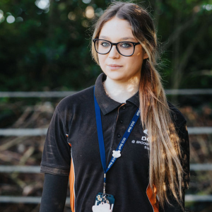 Person with long brown hair wearing a black jumper in a field with a gate.
