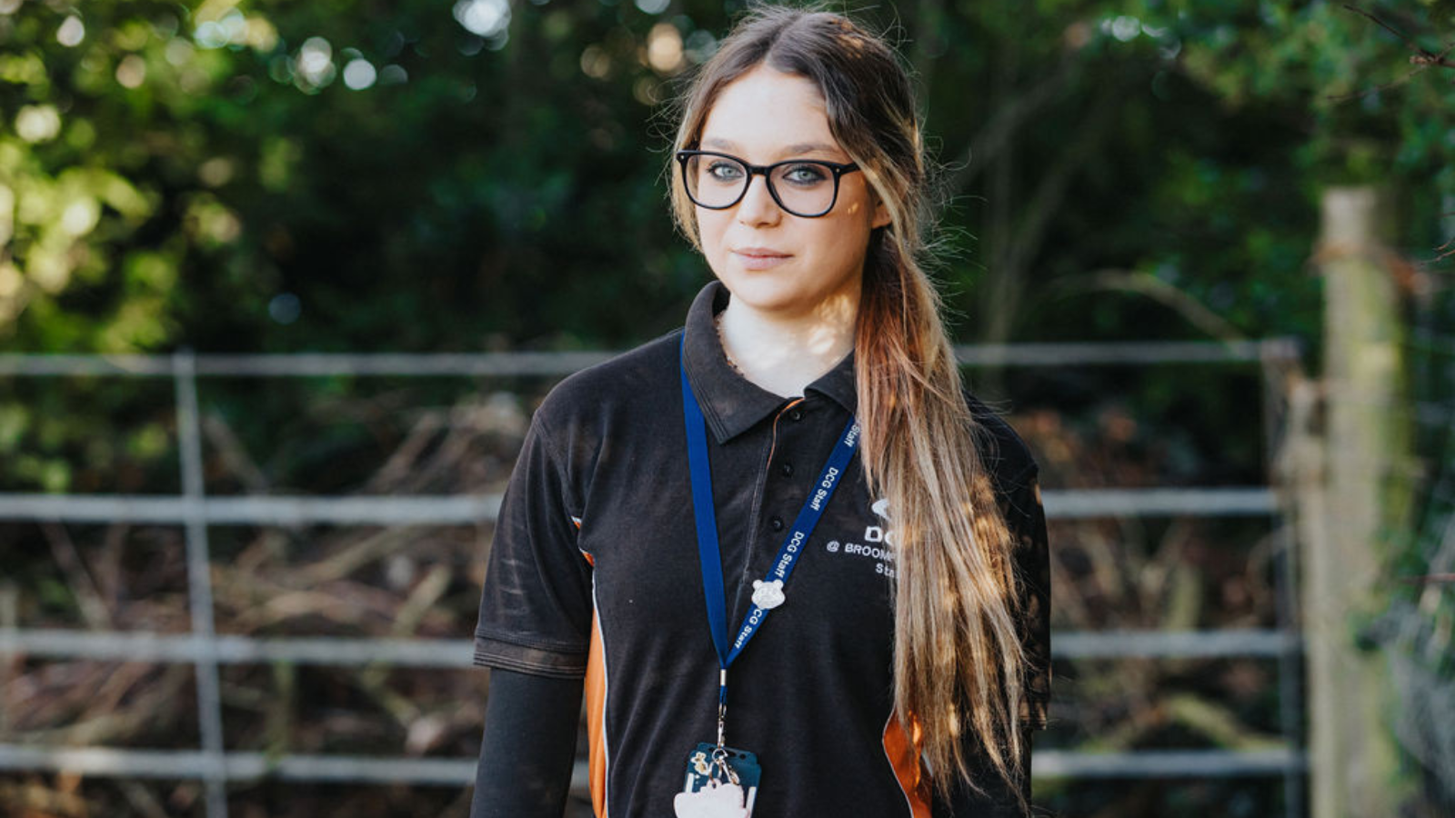 Person with long brown hair wearing a black jumper in a field with a gate.