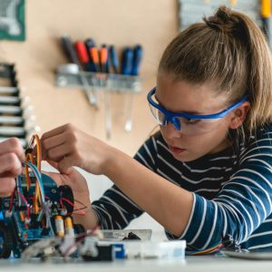 Student assembling a robotics project with wires and electronic components on a workbench in a STEM classroom. Tools and equipment are organised on a pegboard in the background, highlighting practical engineering skills taught in technical courses.