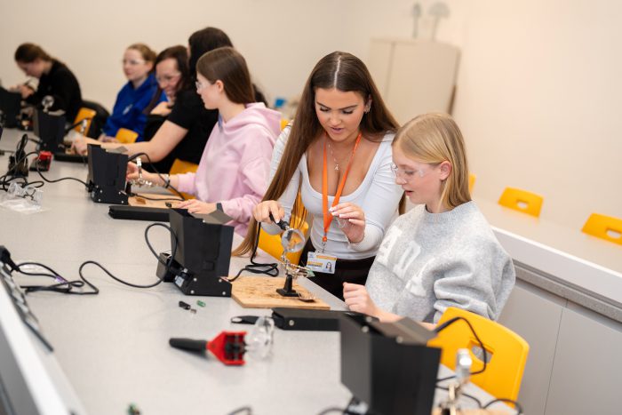 A woman in engineering guides a student through a practical STEM activity at a workstation, helping her use soldering equipment in a classroom setting.