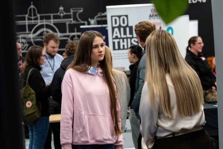 Students at a STEM and engineering event talk near industry exhibition stands, surrounded by engineering‑themed displays and informational banners.