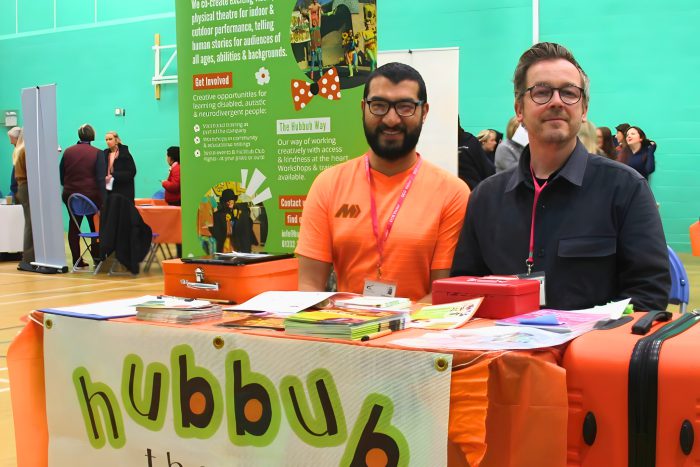 Two representatives from Hubbub Theatre Company sit behind an orange‑themed stall displaying resources and promotional materials at the Inclusion Showcase 2026.