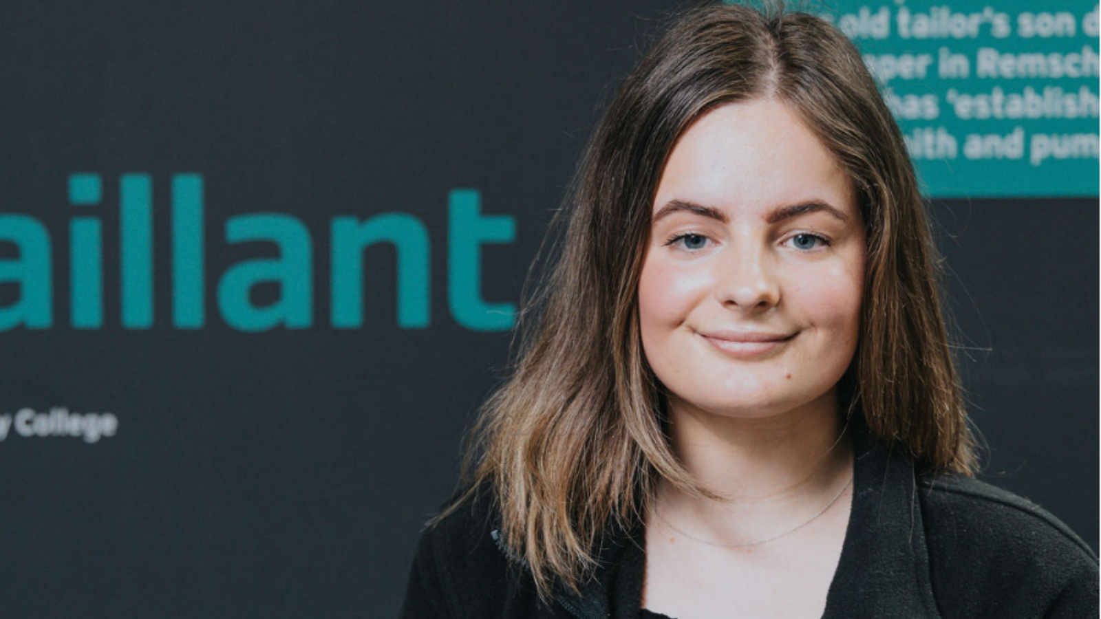 Former Derby College T Level student Grace, wearing a black fleece, stood in front of a wall that reads 'Vailant'.