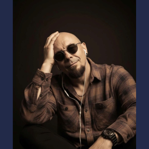 Derby College Distance learning student wearing a checked shirt and sunglasses against a dark background.