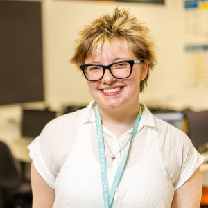 Digital multimedia student standing in a college classroom with computers, representing an Extended Diploma success story, with desks, monitors and educational posters visible in the background, highlighting creative media study and progression