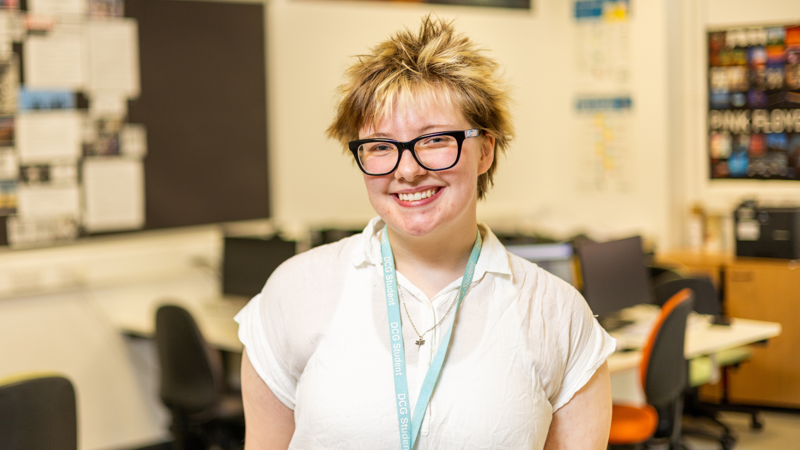 Digital multimedia student standing in a college classroom with computers, representing an Extended Diploma success story, with desks, monitors and educational posters visible in the background, highlighting creative media study and progression