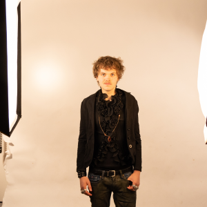 Fashion student from Derby College modelling an all-black outfit in a studio, inspired by Vivienne Westwood and urban rap style, standing between professional photography lights.