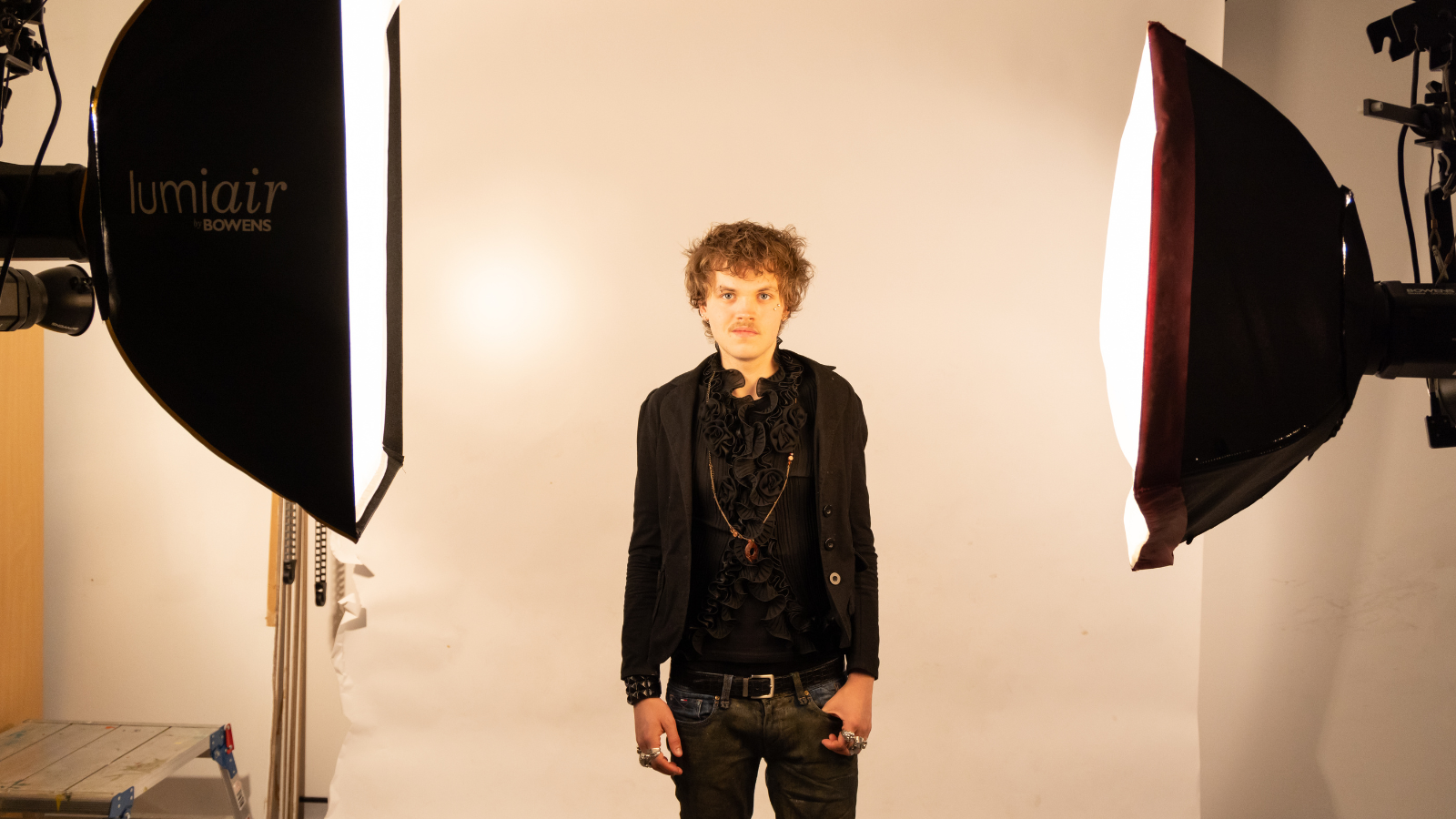 Fashion student from Derby College modelling an all-black outfit in a studio, inspired by Vivienne Westwood and urban rap style, standing between professional photography lights.