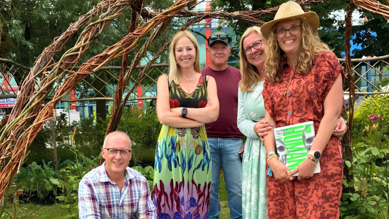 Five Derby College Horticulture students posing together in a garden, standing and crouching beneath a woven willow arch, with greenery and plants surrounding a mosaic feature on the ground in a landscaped outdoor creative space that won silver at Gardner's World Live